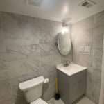 Modern bathroom with marble tile walls, vanity sink, toilet, and round mirror under light fixture