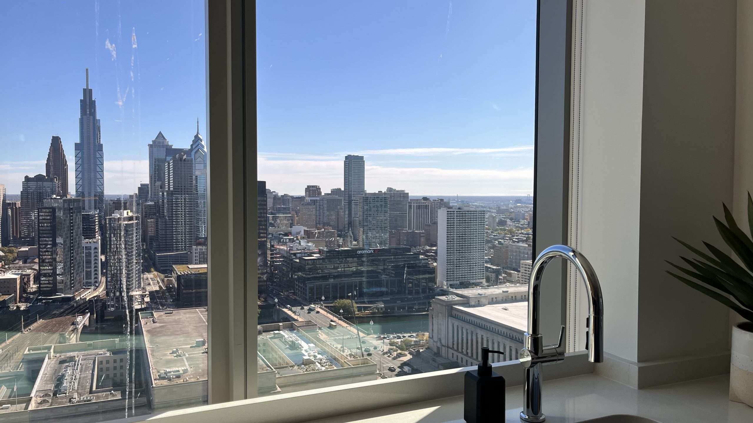 View from modern kitchen with stainless steel sink and faucet through large window overlooking a city skyline with skyscrapers